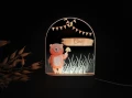 Personalizowana lampka LED z motywem misia na tle wysokiej trawy, trzymającego grzyba, ozdobiona pastelowymi girlandami, idealna do pokoju dziecka.