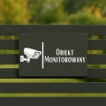 Czarna tabliczka z napisem „Obiekt monitorowany” na ogrodzeniu.
