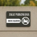 Czarna tabliczka z białym napisem o zakazie parkowania zamocowana na płocie.